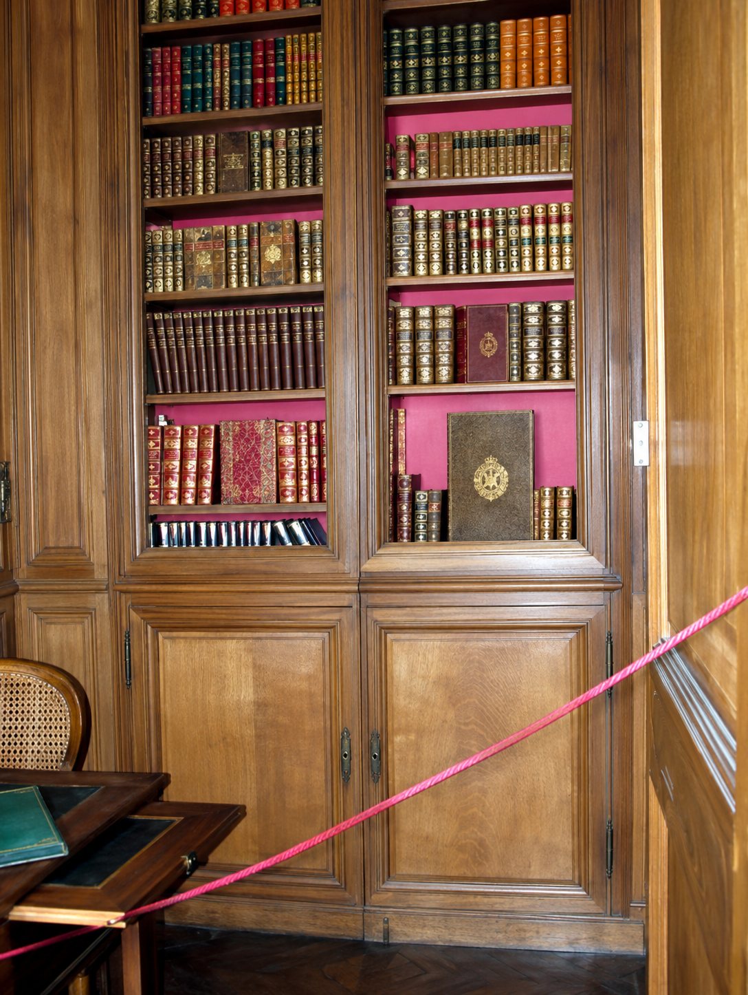 Une grande bibliothèque ancienne en bois sombre remplie de livres reliés, protégée par un ruban rose tendu devant elle dans une pièce historique.