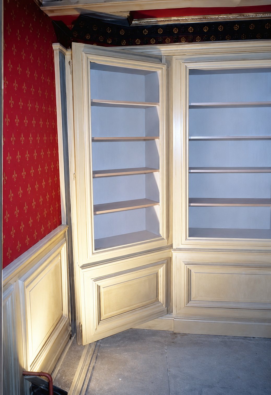 Une bibliothèque d'angle en bois patiné clair, avec étagères vides et lambris, installée dans une pièce aux murs rouges à motifs.