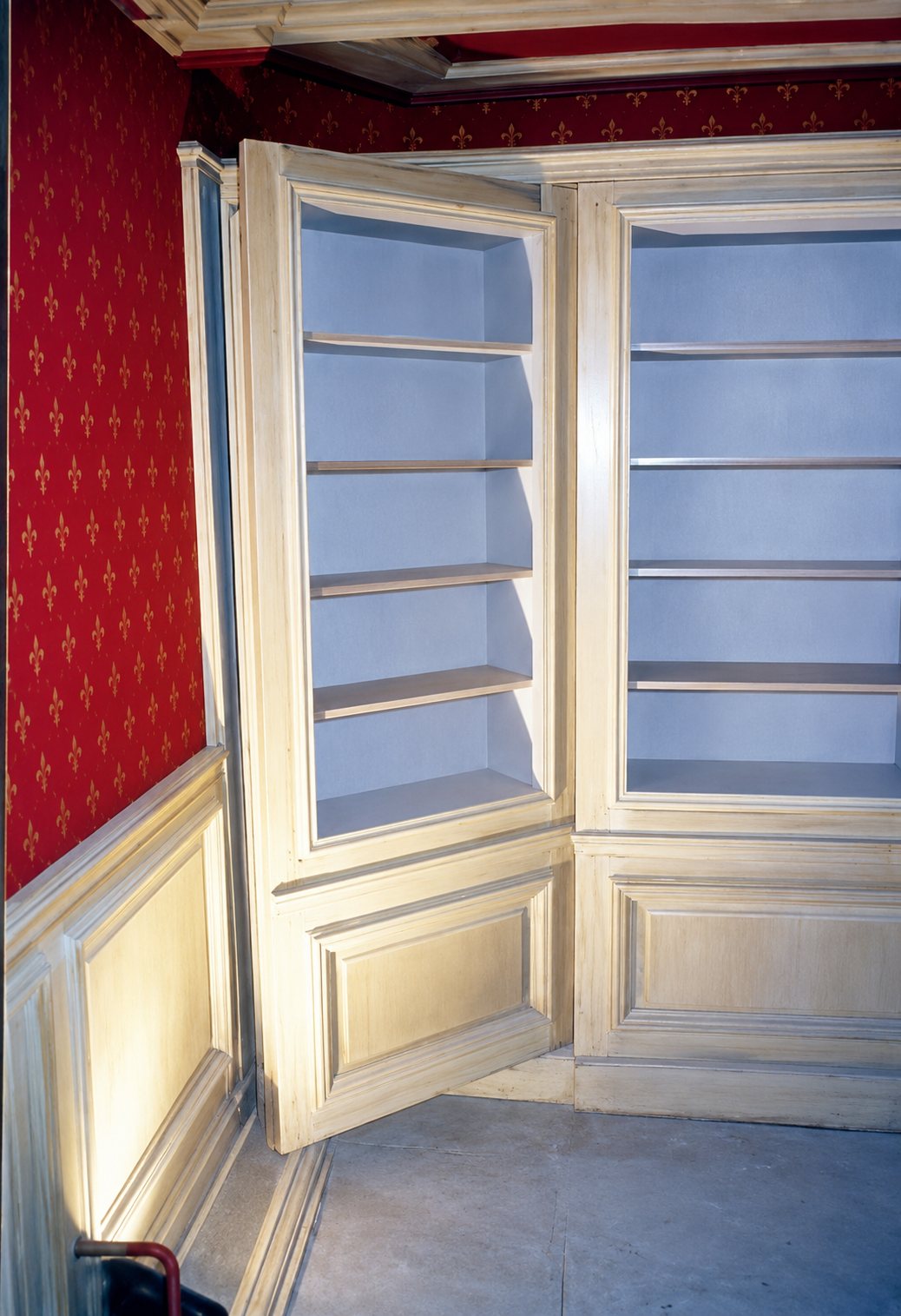 Bibliothèque d'angle en bois clair patiné avec étagères ouvertes et lambris, dans une pièce aux murs rouges à motifs.