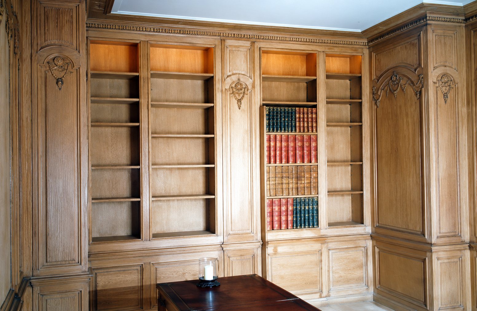 Une pièce aux boiseries en bois clair sculpté comprend de grandes étagères de bibliothèque encastrées, garnies de quelques livres reliés.