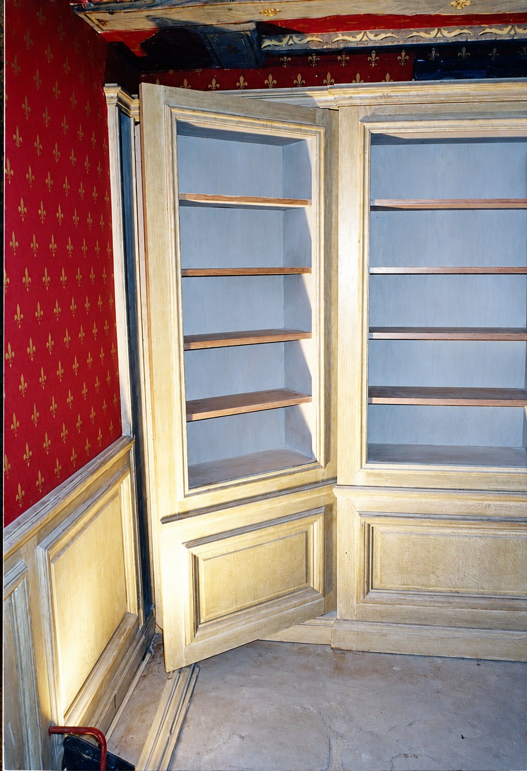Bibliothèque d'angle en chêne clair avec porte ouverte et lambris, devant un mur tapissé de rouge à motifs de fleurs de lys.
