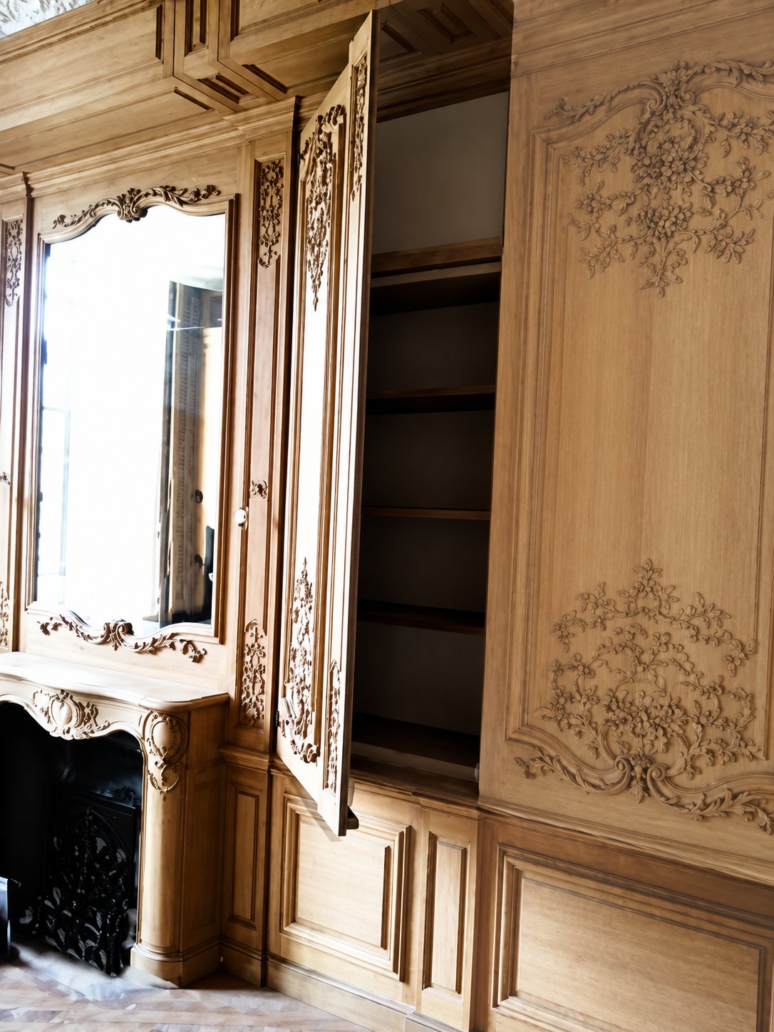 Intérieur d'une pièce habillée de boiseries en bois clair aux motifs sculptés, avec un miroir, une cheminée en marbre sombre et une porte de placard ouverte.