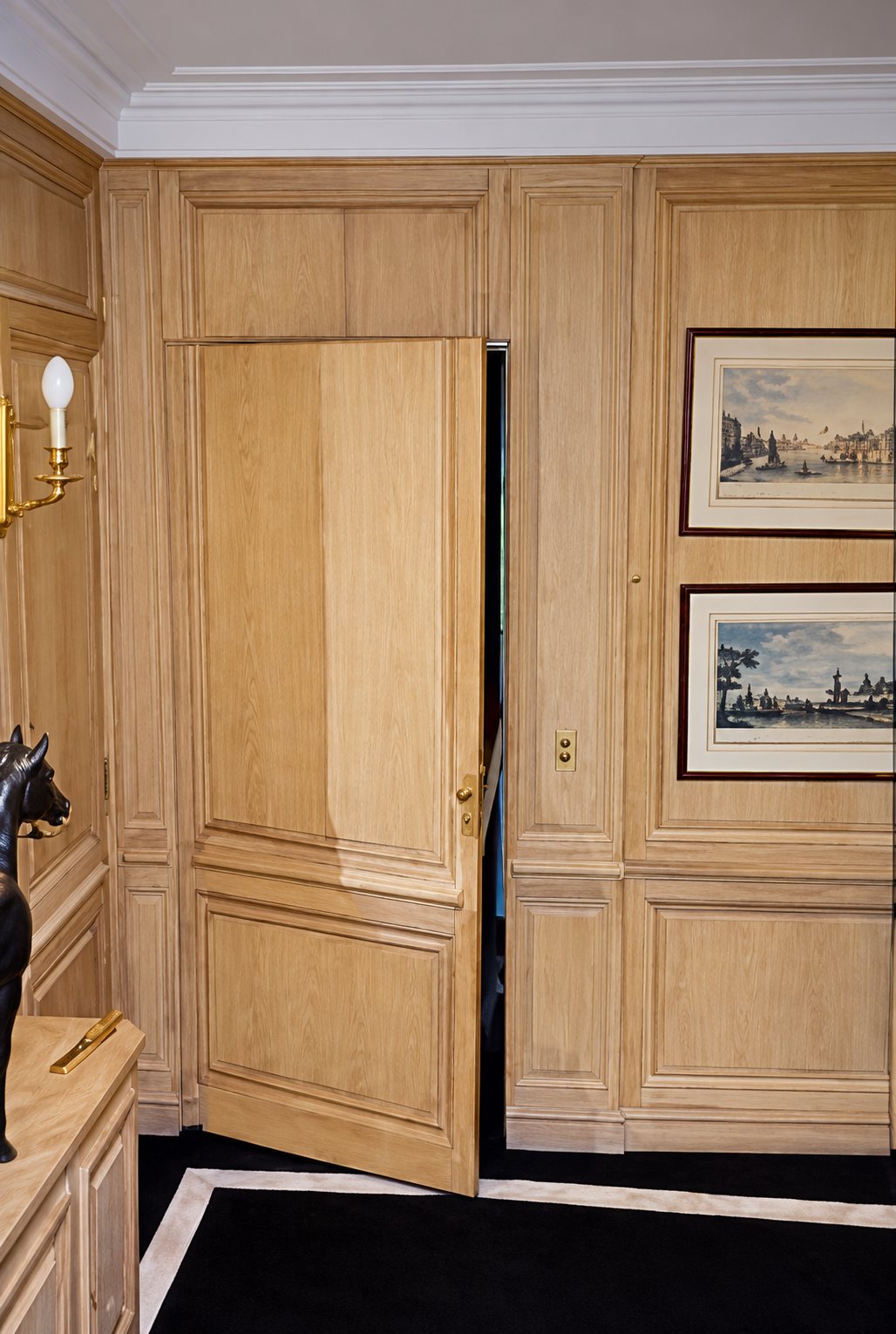 Une pièce aux murs couverts de boiseries en bois clair, avec une porte dérobée entrouverte et deux gravures encadrées au mur.