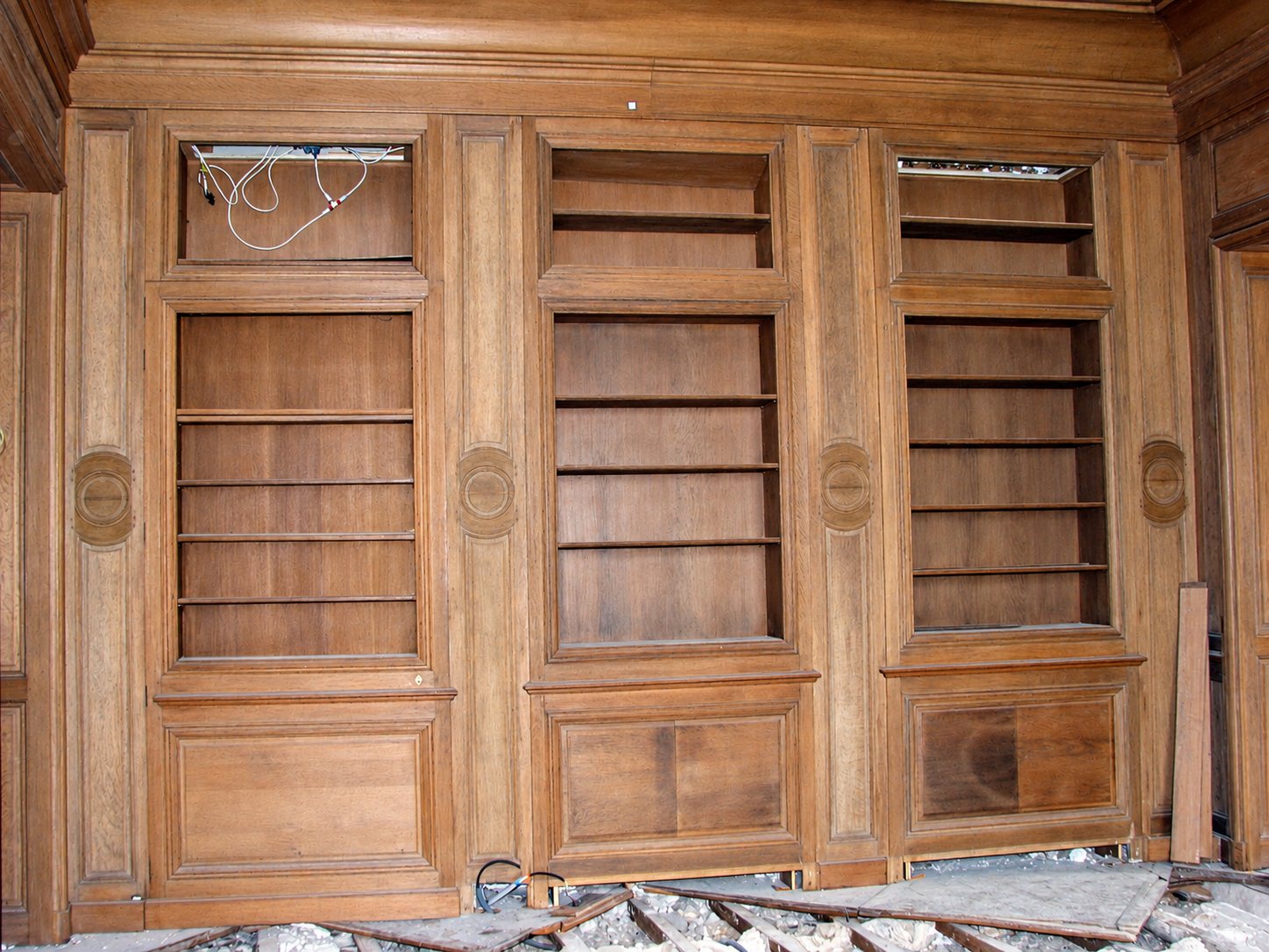 Une grande boiserie ancienne en chêne comporte trois bibliothèques encastrées vides, dans une pièce en travaux dont le sol est jonché de gravats.