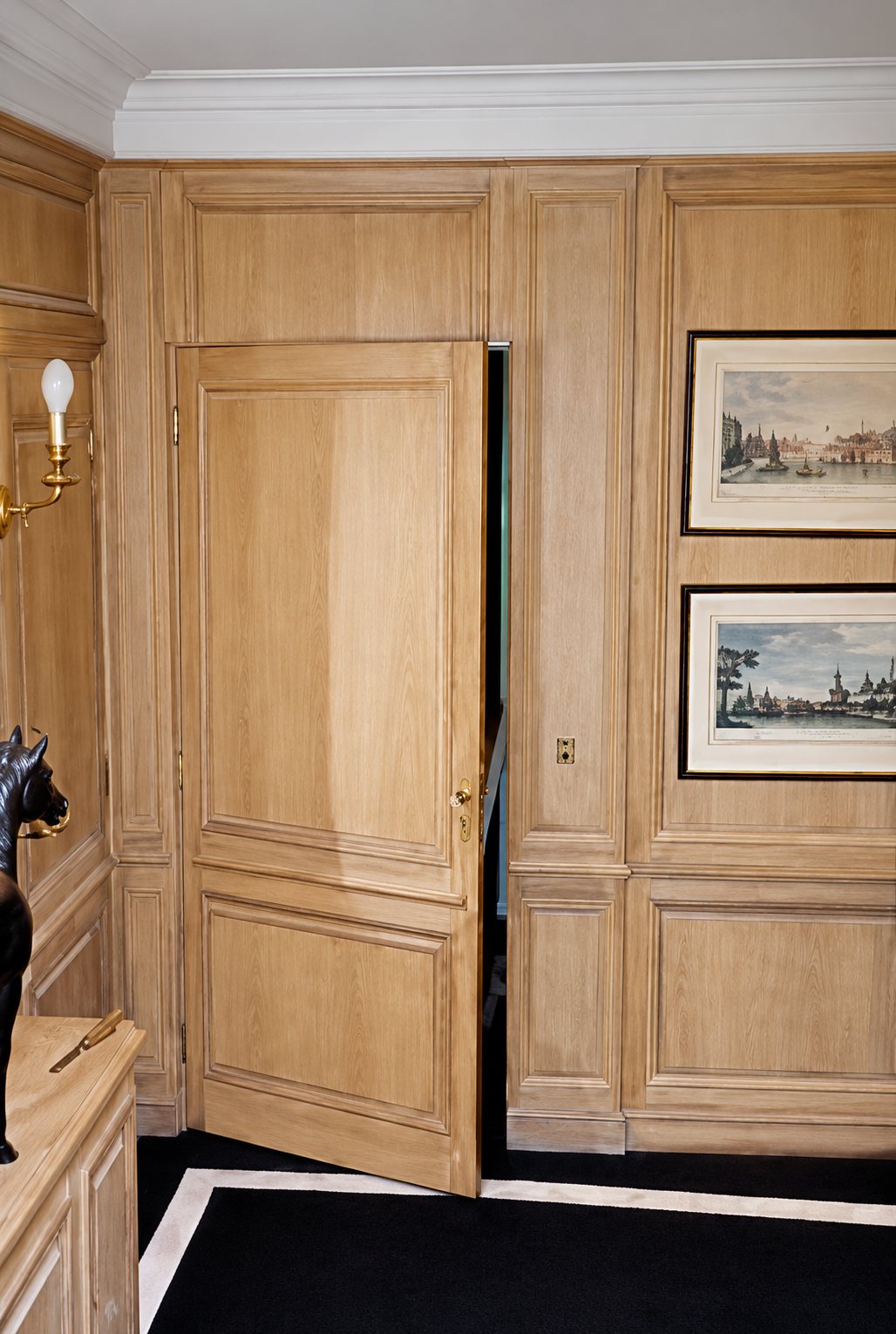 Une pièce aux boiseries en chêne clair dont une porte secrète est entrouverte, avec deux gravures encadrées accrochées au mur de droite.