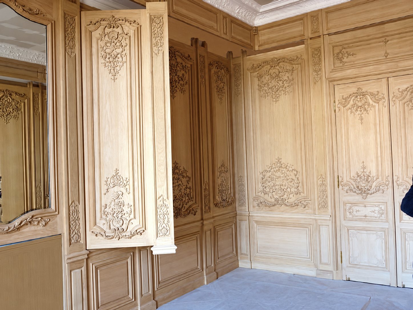 Pièce aux murs couverts de boiseries en chêne clair sculptées de motifs rocaille, avec un miroir à gauche et des portes à double battant à droite.