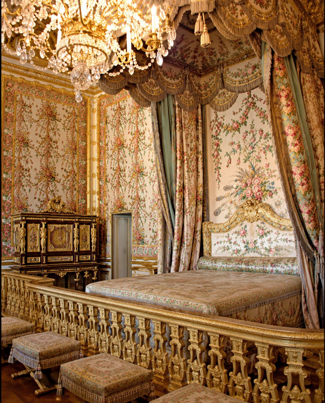 Une chambre historique aux murs ornés de motifs dorés, avec un lit à baldaquin derrière une balustrade sculptée et un lustre en cristal au plafond.