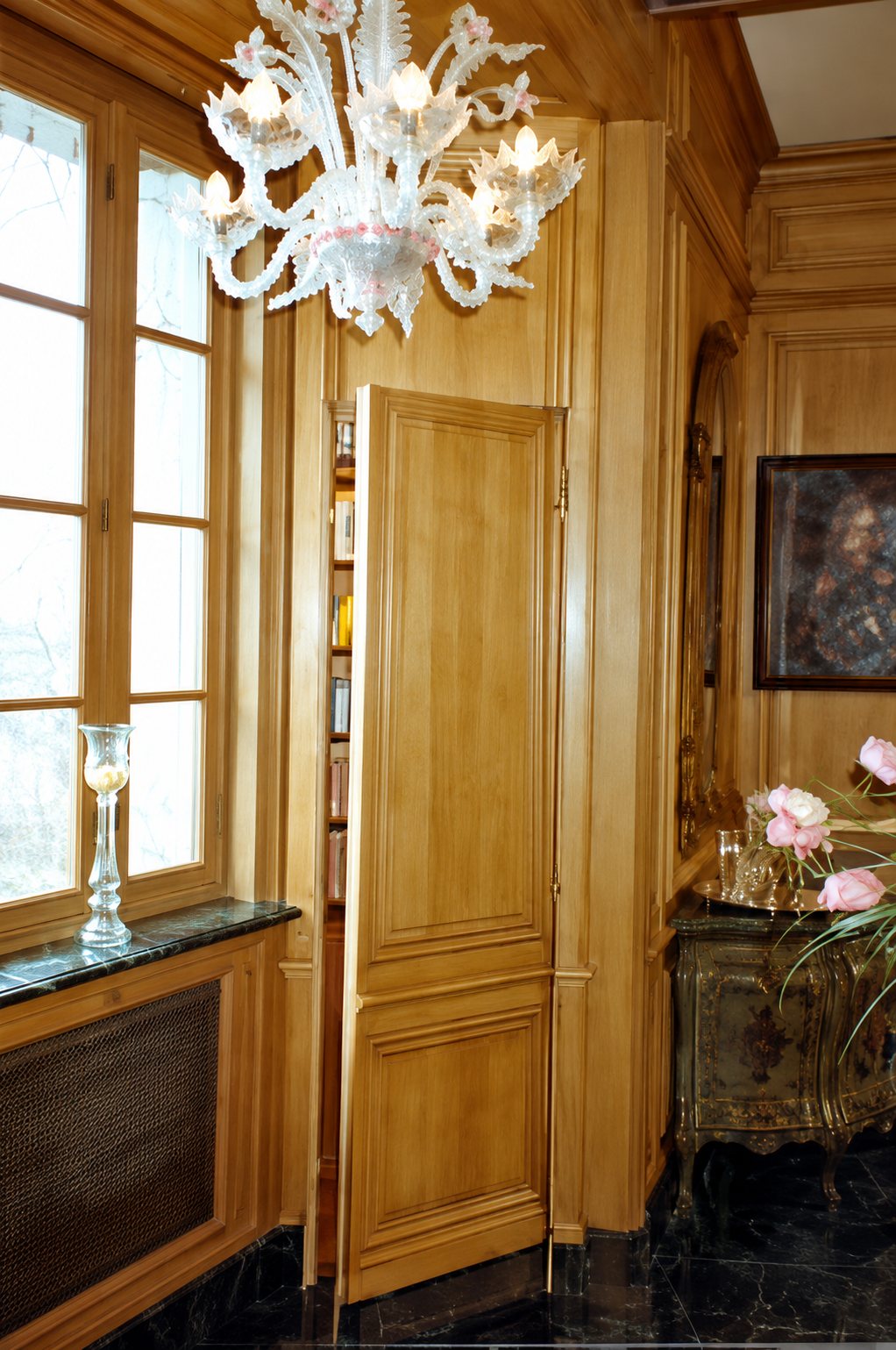 Une porte dérobée en bois clair est entrouverte dans une boiserie, à côté d'une fenêtre, sous un lustre en verre de Murano.