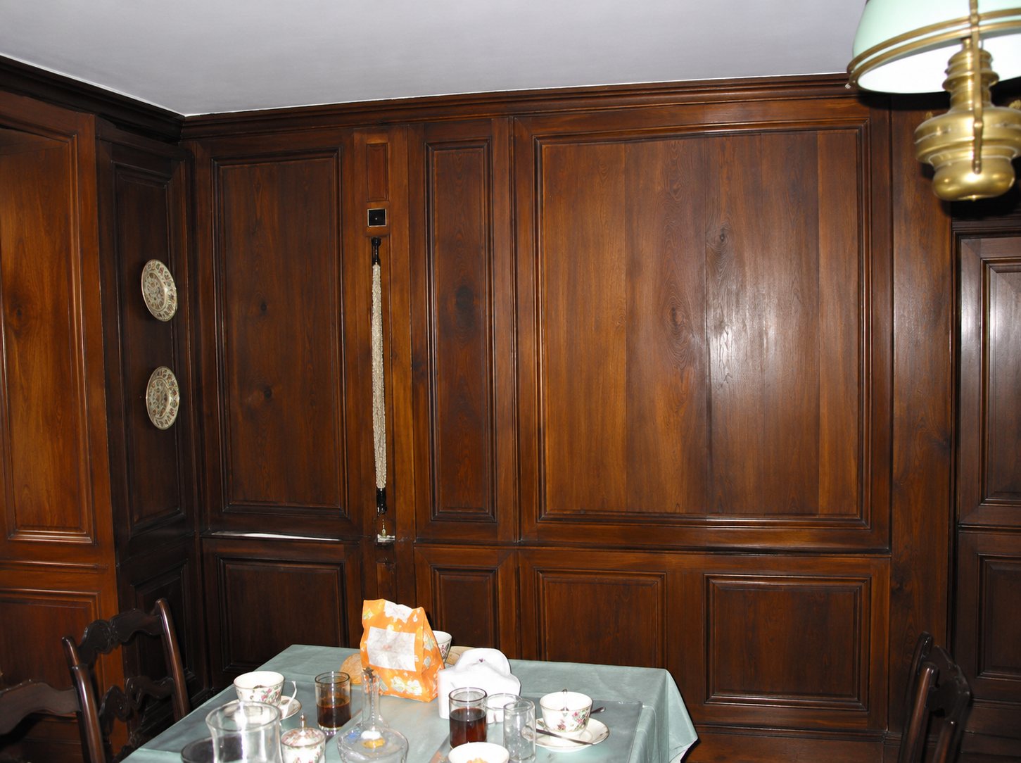 Une salle à manger aux murs entièrement recouverts de boiseries en bois sombre, avec une table dressée au premier plan et une lampe suspendue à droite.