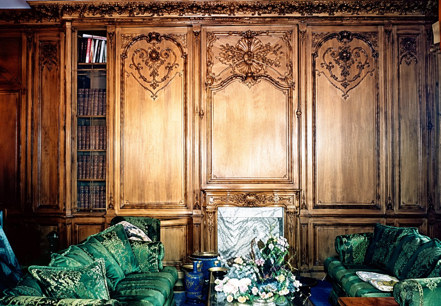 Un salon aux grandes boiseries en bois sculpté de style classique, meublé de fauteuils et canapés en tissu vert devant une cheminée.