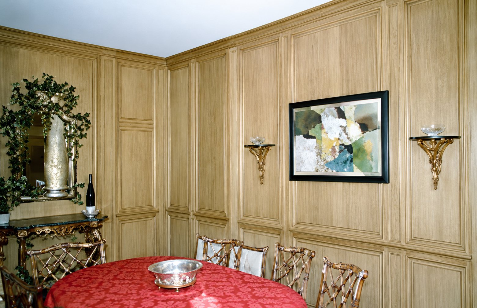 Pièce aux murs couverts de boiseries en bois clair, avec un tableau encadré, deux appliques, un grand miroir ancien et une table ronde recouverte d'une nappe rouge.