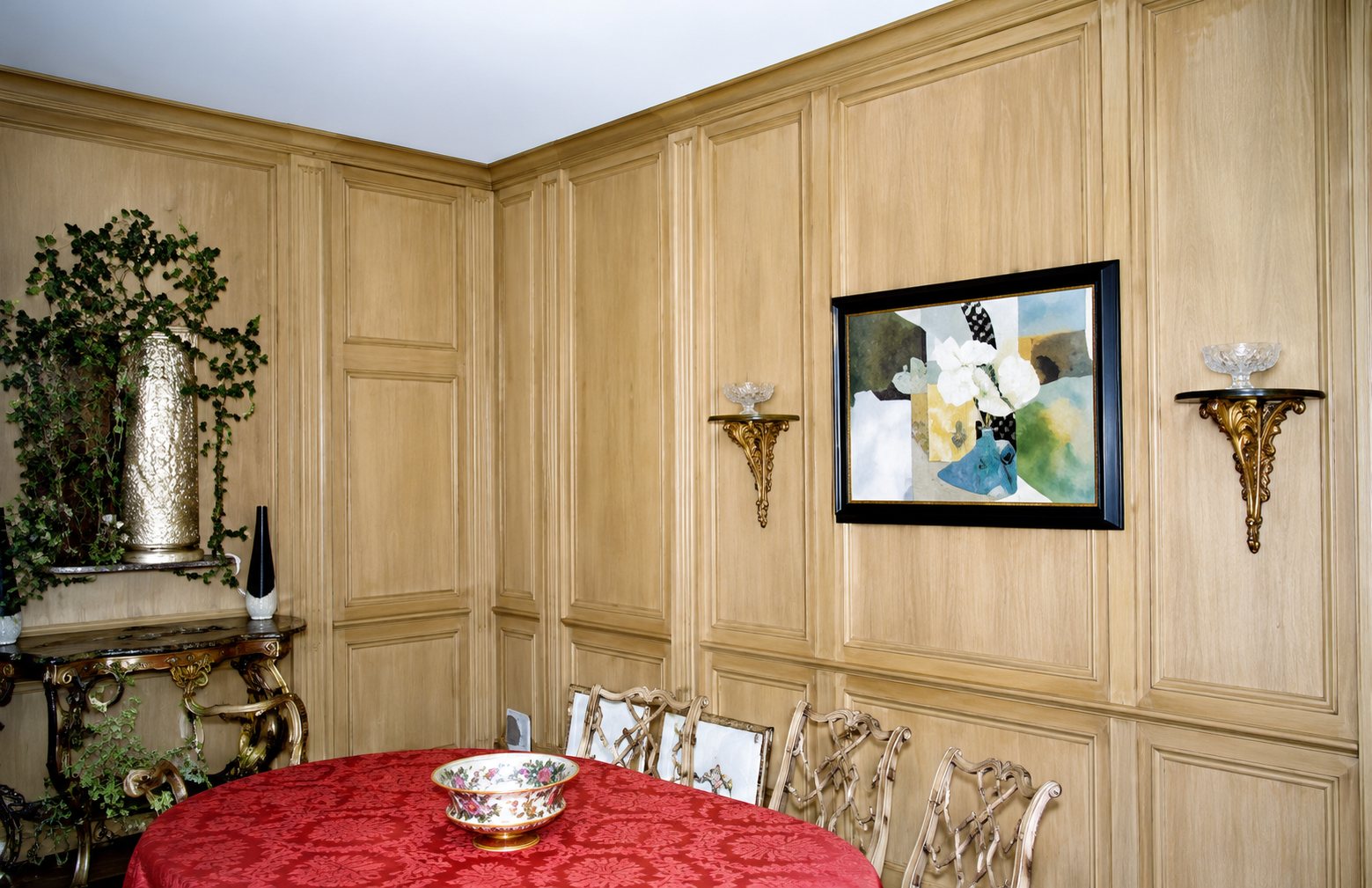 Pièce aux murs recouverts de boiseries claires, avec un tableau encadré, deux appliques, un miroir ancien et une table ronde à nappe rouge.